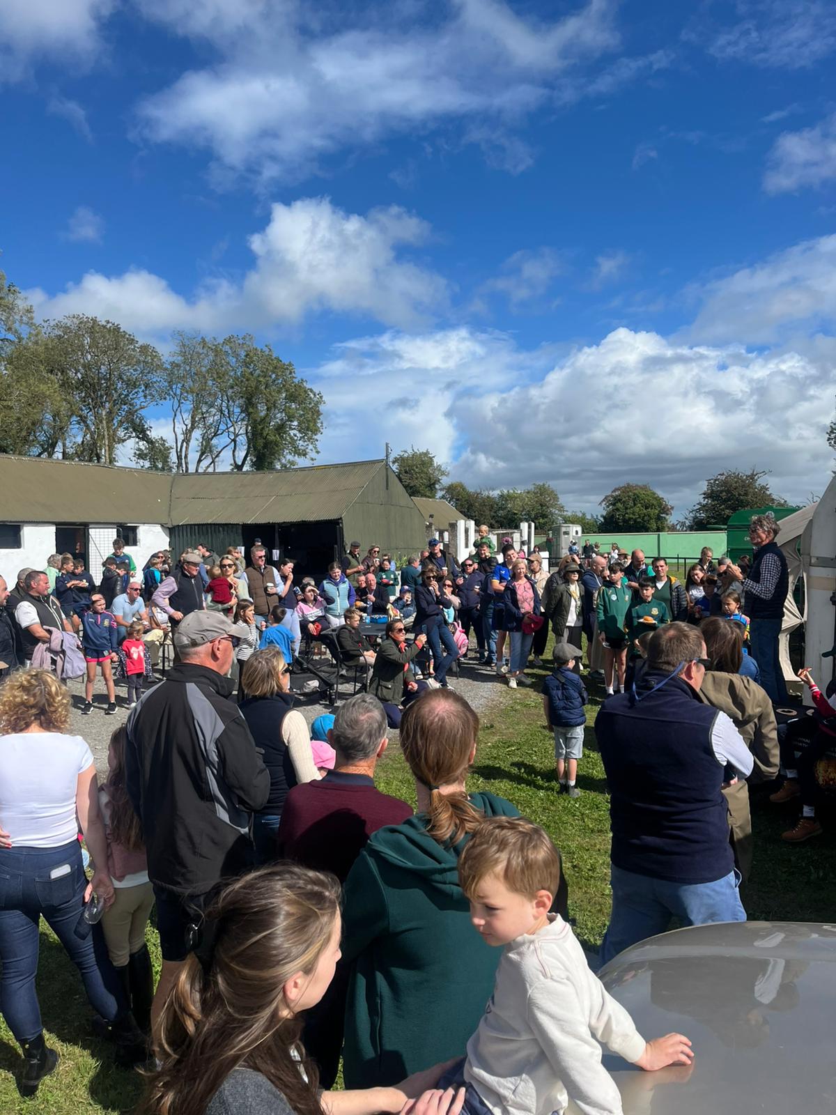 A large gathering of adults and children at an outdoor rural community event under a blue sky